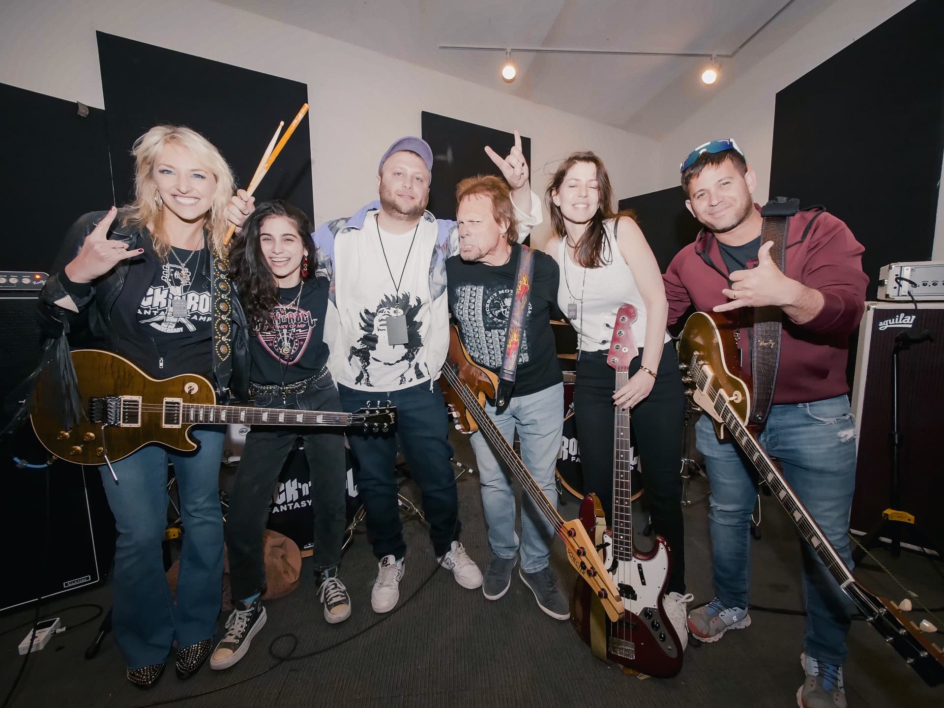 Six musicians pose with guitars and drumsticks in a rehearsal room.
