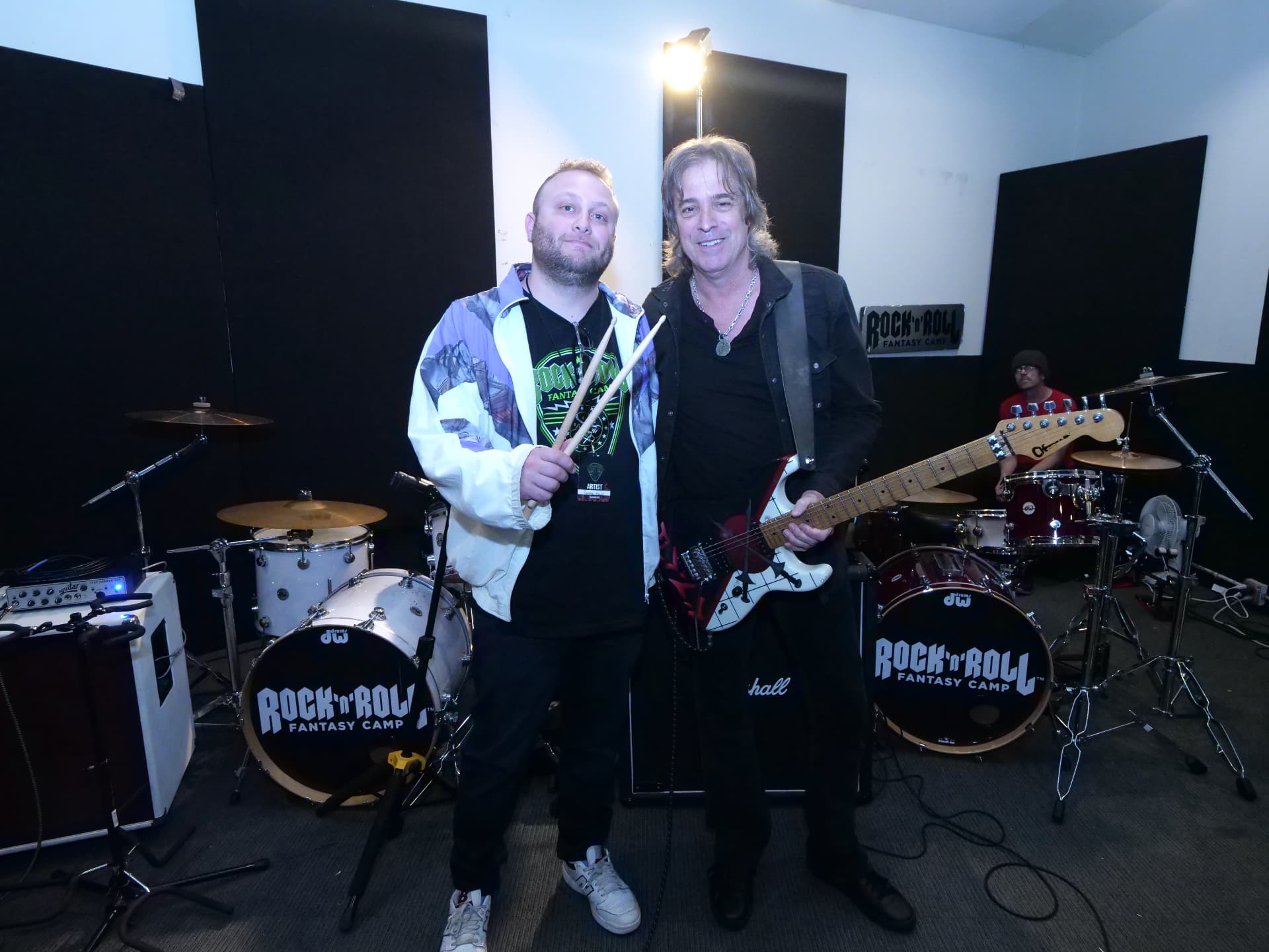 A drummer holding sticks stands beside a guitarist in a Rock 'n' Roll Fantasy Camp rehearsal room.